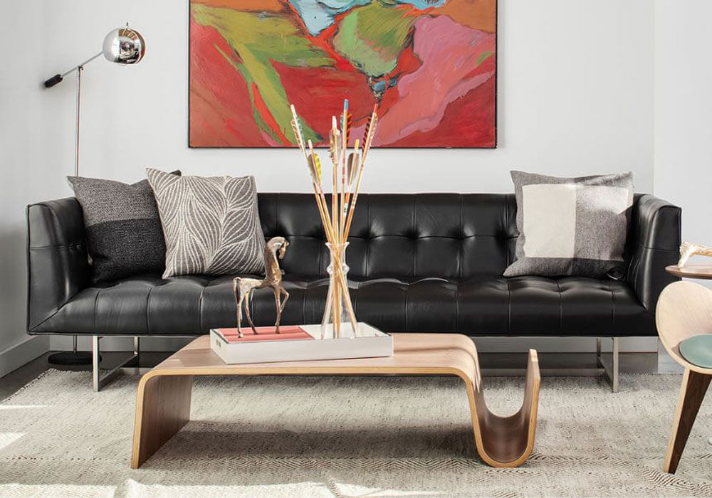 Modern living room featuring a black tufted leather sofa with assorted throw pillows, a contemporary wooden coffee table, and vibrant abstract artwork on the wall. A sleek floor lamp and decorative accents, including a horse figurine and reed diffuser, complete the look.
