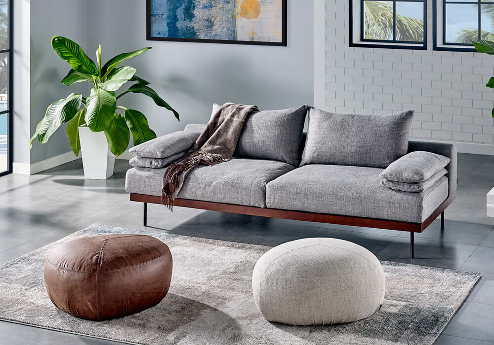A modern living room with a gray sofa adorned with a brown throw blanket, two poufs (one brown leather and one beige fabric) on a soft rug, a large potted plant, and abstract artwork on the wall. Large windows in the background let in natural light.