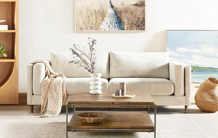 A minimalistic living room features a light beige sofa with a plaid blanket draped over one arm. In front of the sofa is a wooden coffee table with decorative items. A coastal-themed painting hangs above the sofa, and a second painting rests against the wall.