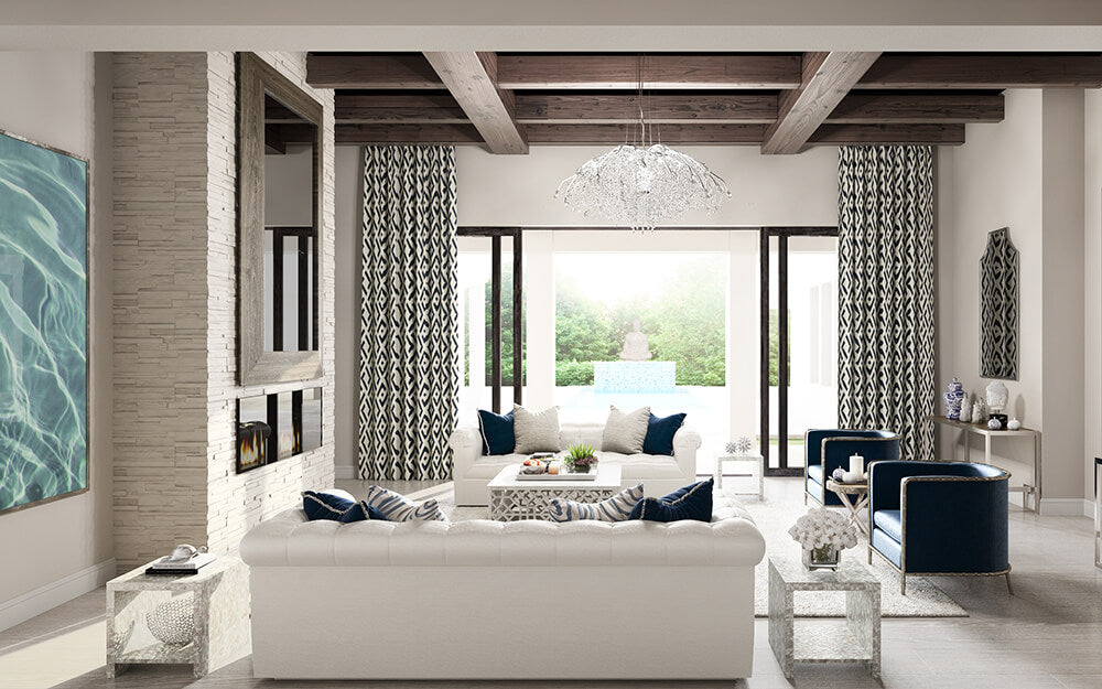 A modern living room with a white sofa, blue armchairs, and a large coffee table. The room features a stone fireplace with a TV above it, wooden ceiling beams, patterned curtains, and a chandelier. Large windows offer a view of a greenery-filled outdoor space.