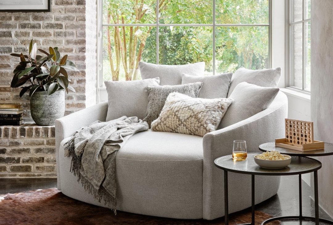 A cozy reading nook features a large, round, light gray armchair adorned with various pillows and a soft throw blanket. A small side table holds a glass of whiskey and a bowl of popcorn. Nearby, a potted plant and a large window with a garden view complete the scene.