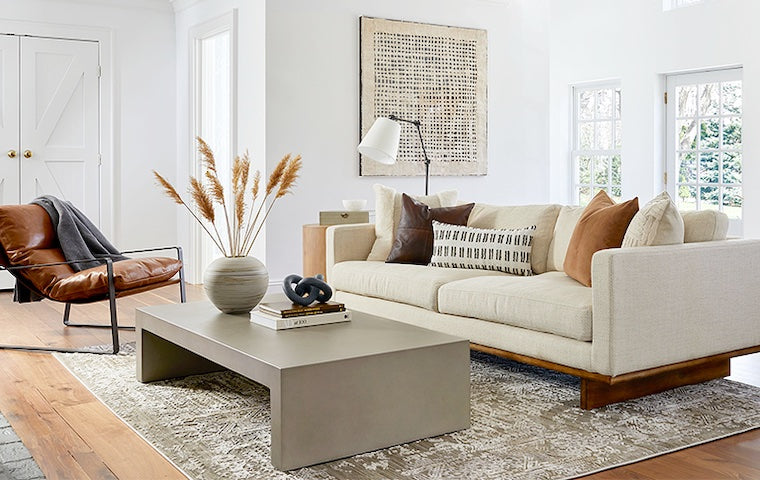 A bright living room features a beige sofa adorned with earth-tone pillows, a modern concrete coffee table with decor items, a leather chair with a metal frame, a patterned area rug, and a large abstract wall art piece. Natural light streams through multiple windows.
