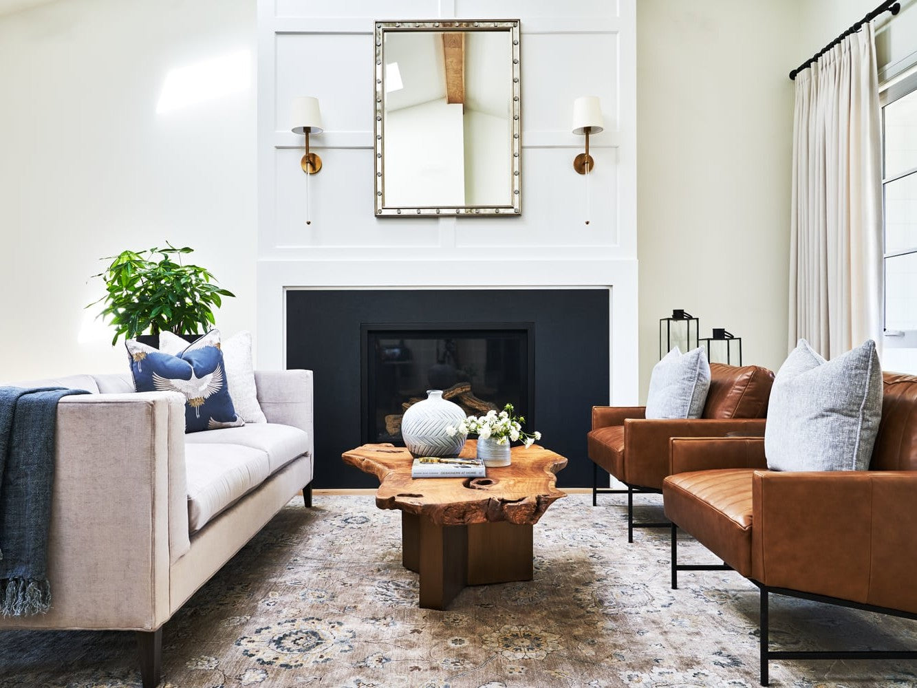 A modern living room features a white sofa with blue and white pillows on the left, facing two brown leather armchairs with gray pillows on the right. A wooden coffee table with decorative items is centered on a patterned rug. A mirror and wall sconces are above a fireplace.