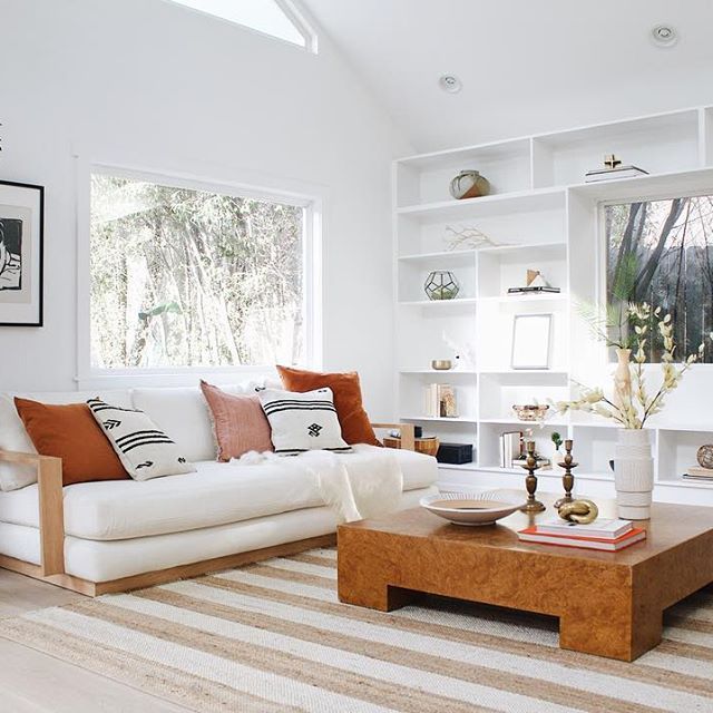 A bright, modern living room with a white sofa adorned with brown and white patterned pillows. A wooden coffee table sits atop a striped rug. Built-in shelves with decorative items are against the back wall, and a large window allows natural light to flood the room.