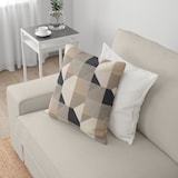 A cozy beige KIVIK Sofa featuring two cushions, one displaying a geometric pattern in neutral tones, rests invitingly. Beside the sofa, a small white side table accommodates a plant and two teacups. White curtains are drawn open, bathing the room in natural light.