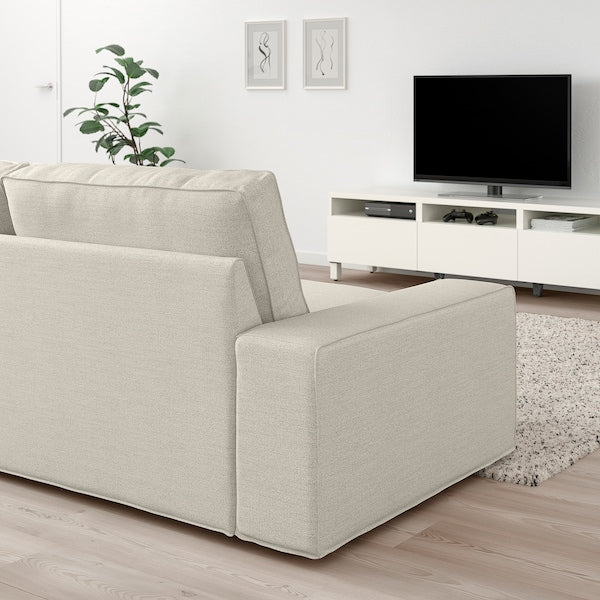 A modern living room features a KIVIK Sofa with Chaise in light gray positioned to face a TV on a white stand. A plant rests in the corner, while two minimalist art pieces adorn the wall. The floor is wooden, complemented by a light-colored rug.