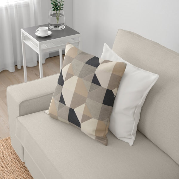 In a well-lit room, the beige KIVIK Sofa with Chaise is adorned with two decorative pillows. One pillow showcases a geometric pattern in neutral tones, while the other is plain white. A small side table next to the sofa holds a potted plant along with a white cup and saucer.