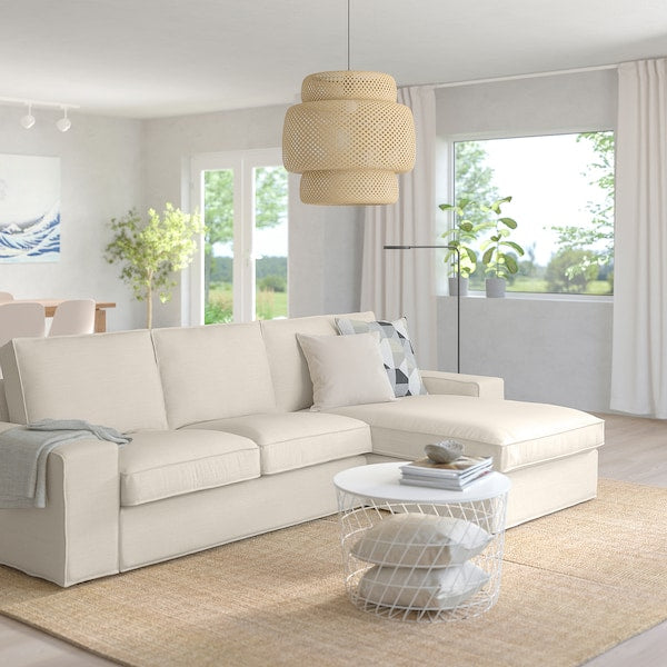 A spacious living room showcasing the KIVIK Sofa with Chaise in light beige, adorned with cushions, alongside a modern round coffee table and a woven pendant light. The large windows provide views of the greenery outside, while a dining area is visible in the background. Natural light floods the space.