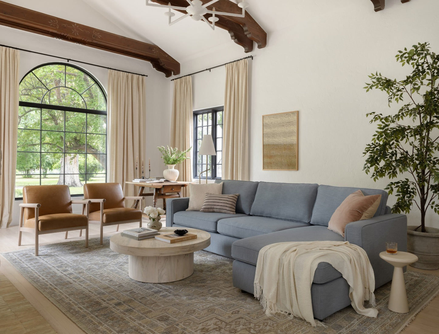 A cozy living room featuring the James 3 Seat Left Chaise 92" Sectional in blue, accompanied by a wooden coffee table and two brown armchairs. The space is accentuated by a large arched window with cream curtains, wooden ceiling beams, and a potted plant. The serene atmosphere is enhanced by the view of greenery outside.