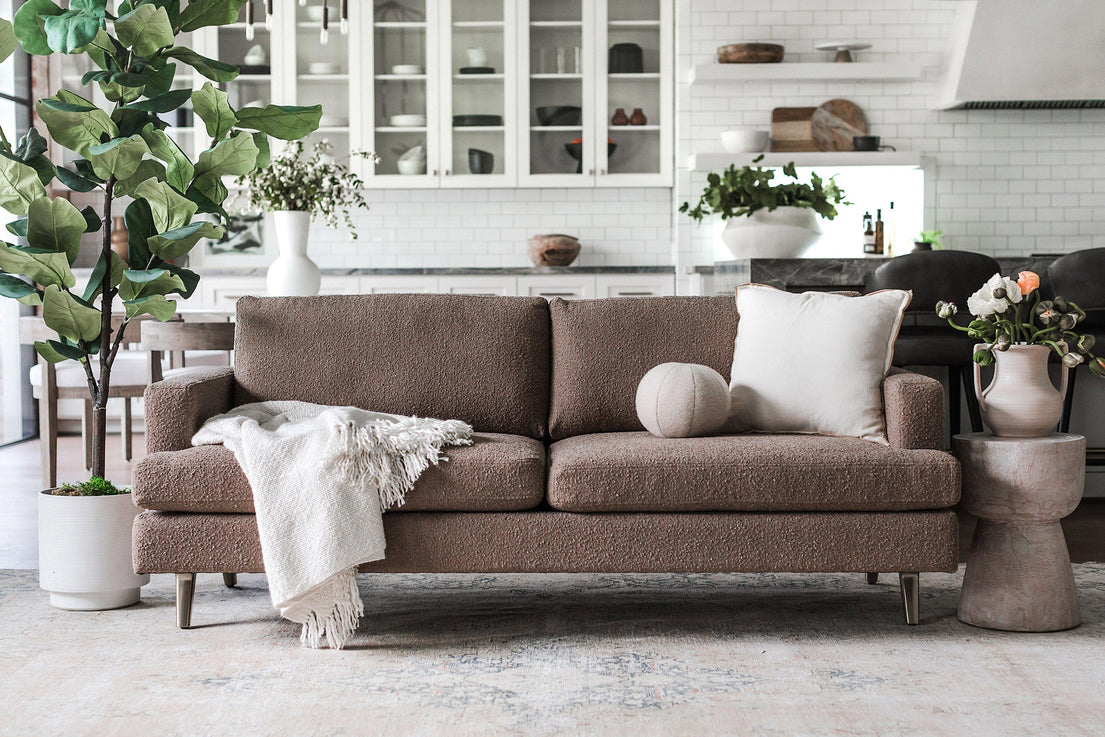 A cozy living room features the Lido Sofa in a warm brown hue, adorned with a white pillow and a throw blanket. A small round cushion adds extra comfort. On either side of the sofa, plants in white pots bring a touch of nature. In the background, you can see a kitchen with pristine white cabinets and countertops.