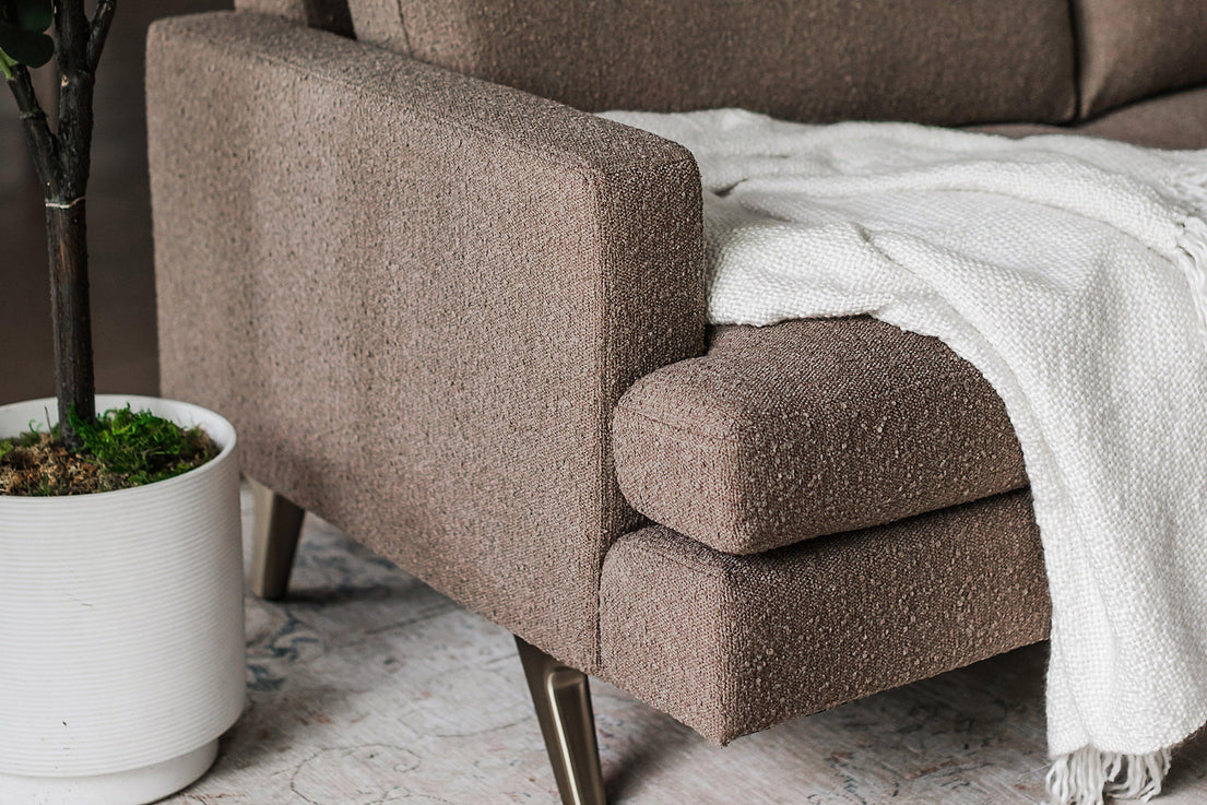 Close-up of the Lido Sofa with a brown textured fabric and sleek metal legs. A soft, white blanket is draped over one arm. A potted plant sits on the floor nearby, adding a touch of greenery to the cozy, modern setting.