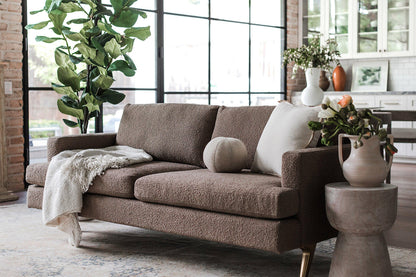 A cozy living room features the Lido Sofa, accented with white pillows and a blanket. A small, round side table is adorned with decorative vases filled with flowers. Beside the sofa stands a large plant, while sunlight streams through an expansive window.