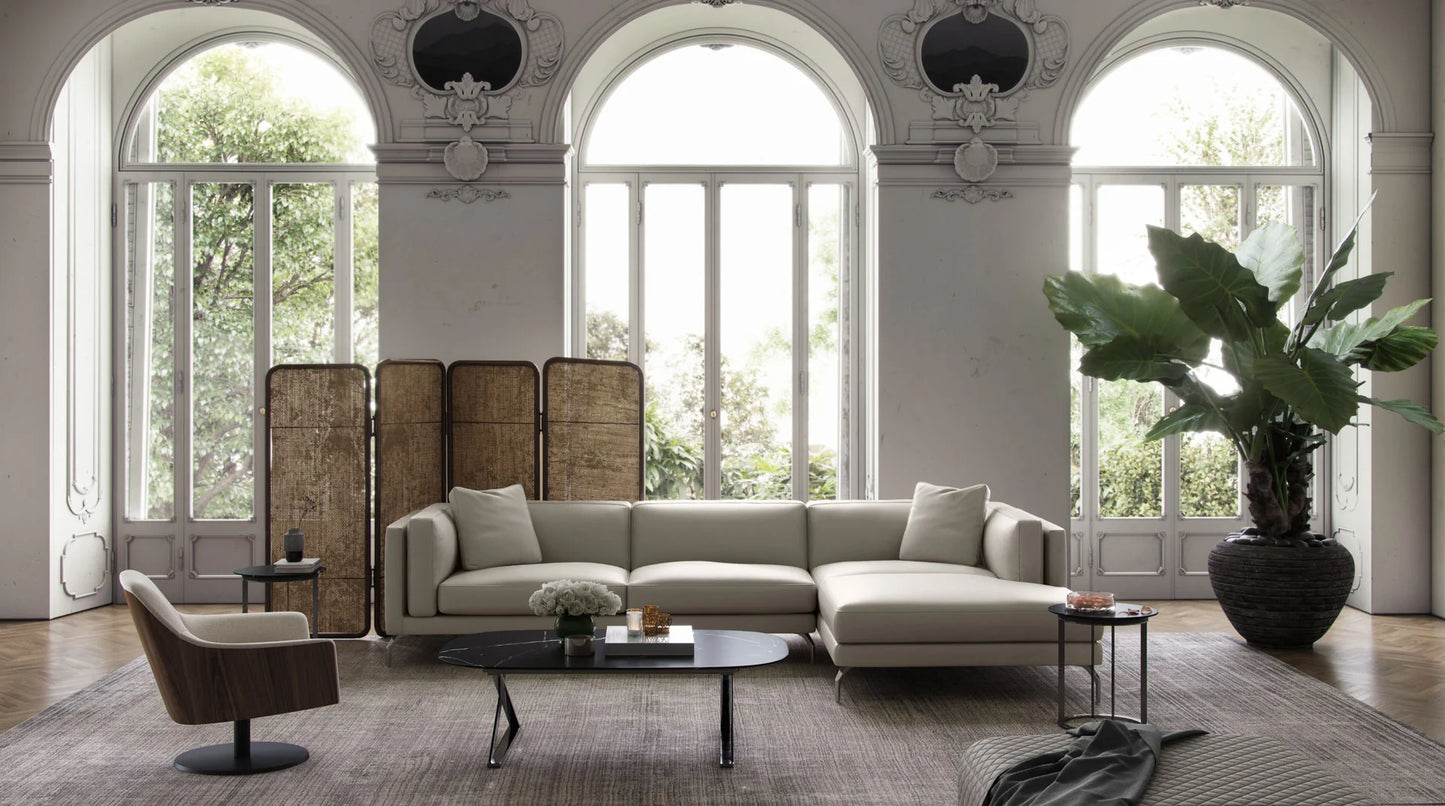 A stylish living room featuring the Reade Sectional Sofa, a round coffee table, two side tables, a wooden chair, and a large potted plant. The room is accented by grand arched windows offering views of greenery outside and includes a wooden room divider.