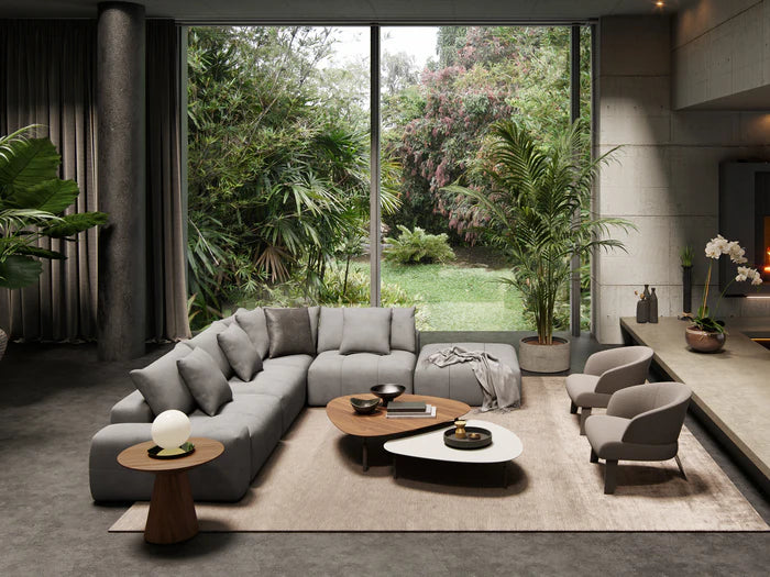 A modern living room featuring the Fullerton Modular Sofa 04 in gray, flanked by two beige armchairs and a pair of wooden coffee tables set on a neutral rug. Large windows offer views of a lush garden outside, while potted plants adorn the interior.