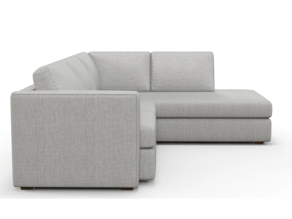 The Lounge Bumper Sectional is a gray L-shaped sofa with clean, minimalist lines. It features plush cushions on the back and seats, making it perfect for a contemporary living space. The sofa rests on low-profile legs, enhancing its modern appearance.