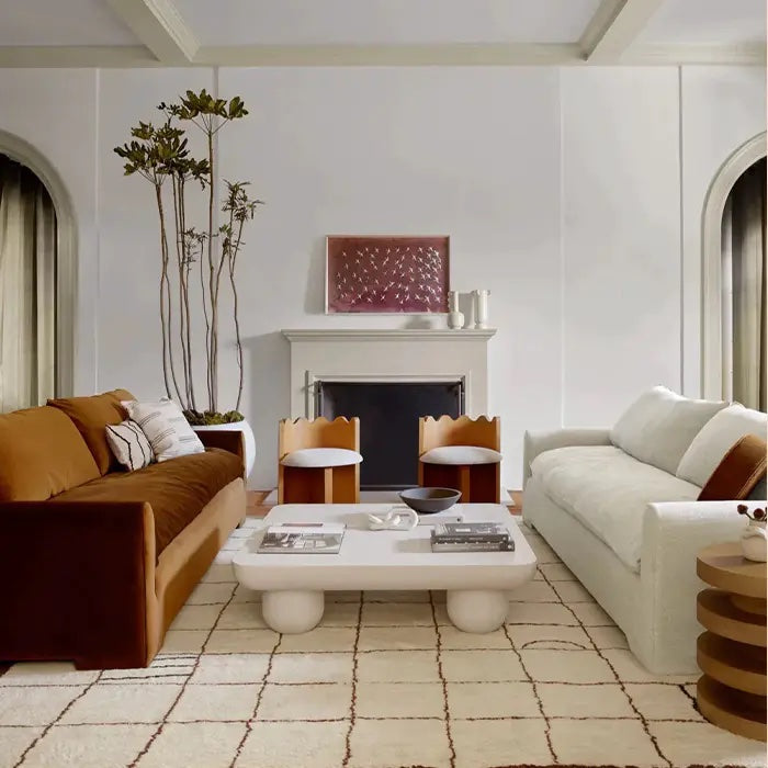 A modern living room with two sofas: one brown and one white, facing each other. There's a white coffee table with decorative items on it, situated on a white rug with a grid pattern. A fireplace with art above it is in the background, flanked by tall plants.