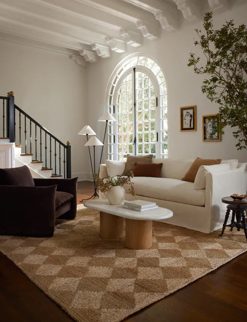 A cozy living room features a white sofa with decorative pillows, a black armchair, a wooden coffee table with vases, and a geometric-patterned rug. A large, arched window with white framing allows natural light to flood the space. A staircase with a black railing is in the background.