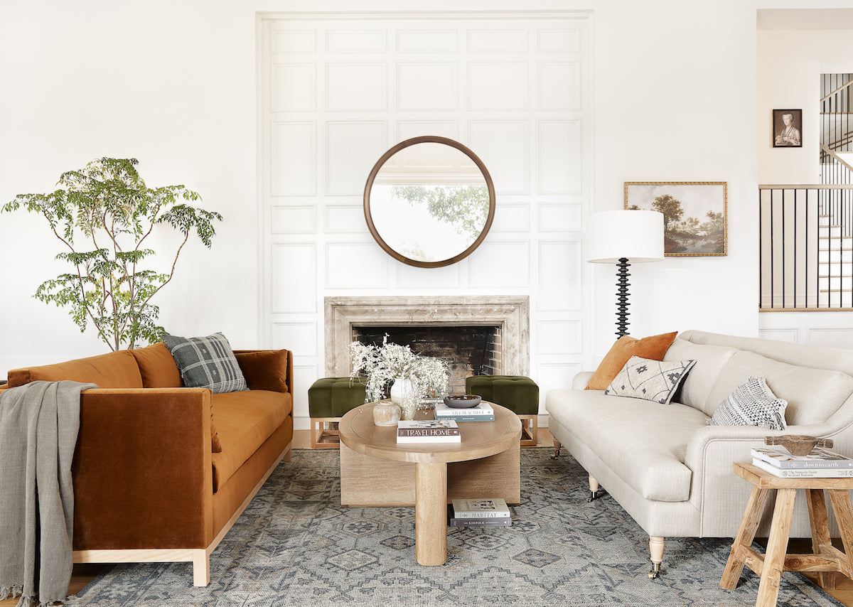 A cozy living room features a mix of modern and rustic decor. It includes an orange velvet sofa, a light beige couch, a round wooden coffee table, and a large round mirror above a fireplace. A green plant and various decorations add warmth to the space.