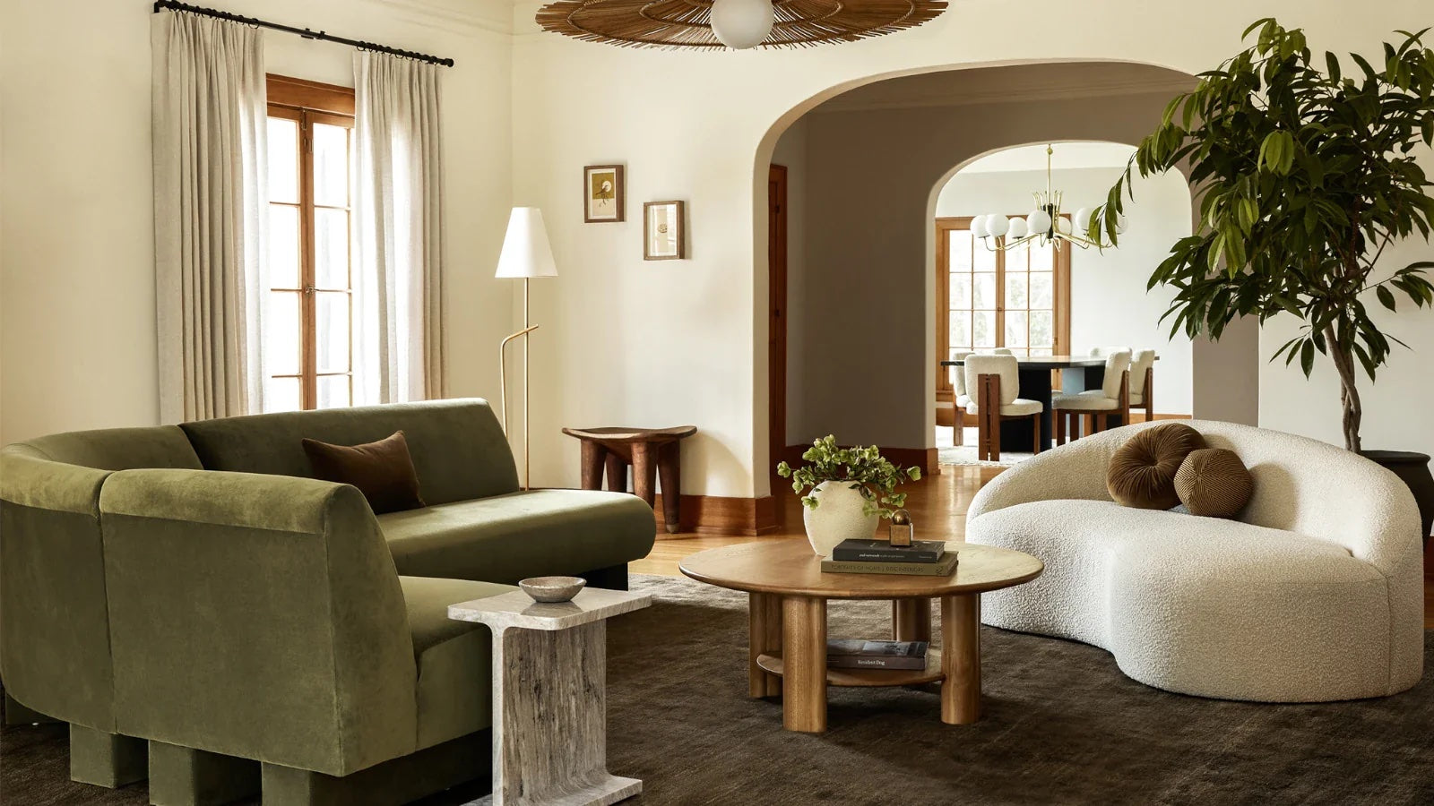 A stylish living area with a green velvet sofa, a white curved sofa, and a round wooden coffee table with decorative items. Large windows with light curtains allow natural light to enter. A potted plant, wall art, and a dining room visible through an arch enhance the decor.