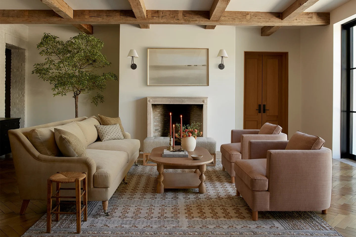 A cozy living room with a beige sofa, two matching armchairs, a wooden coffee table, and a patterned rug. A fireplace adorned with candles and a plant sits between two wall lamps. Wooden ceiling beams and a large window enhance the room's warm and airy ambiance.