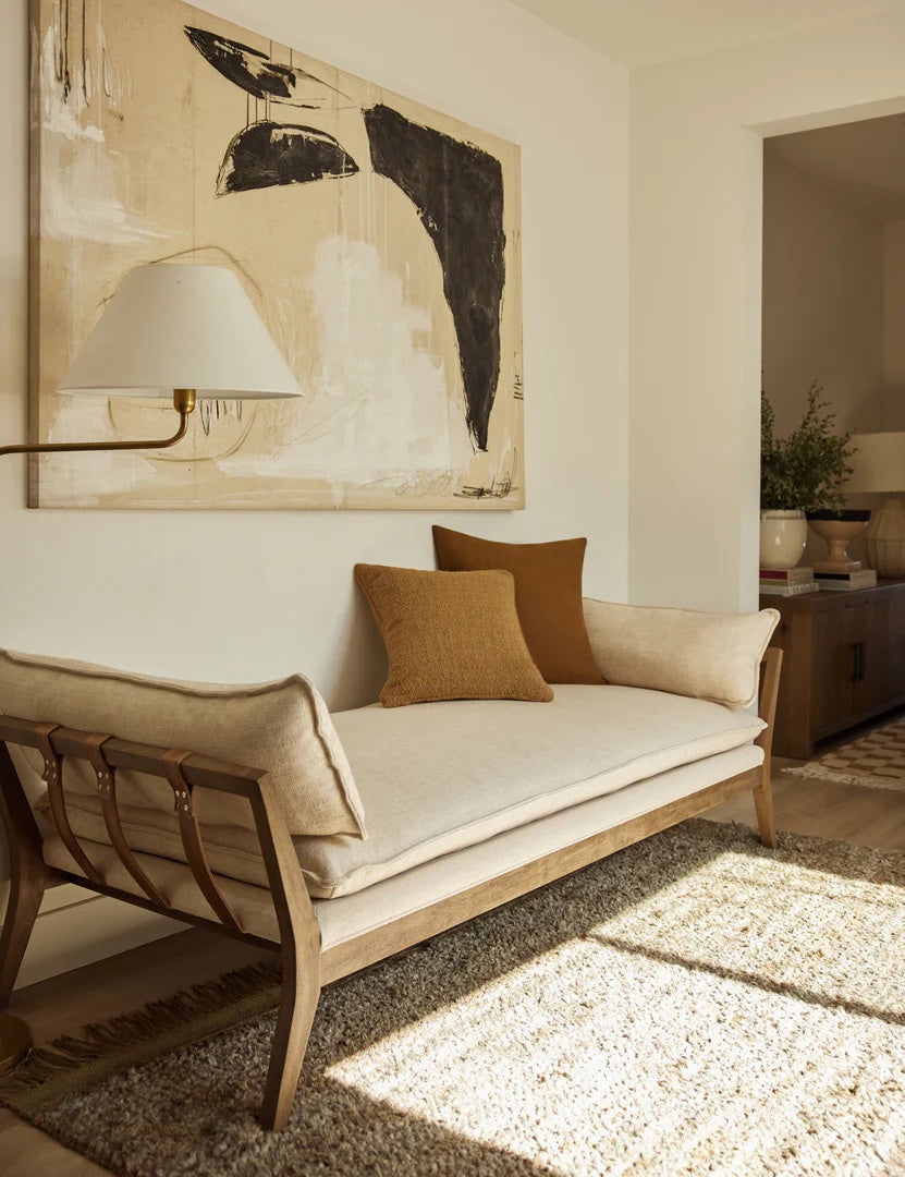 A minimalist living room features a beige wooden-framed daybed with tan and brown cushions. Above it hangs an abstract painting in neutral tones, and a floor lamp with a white shade is mounted on the wall. The floor is covered with a textured beige rug.