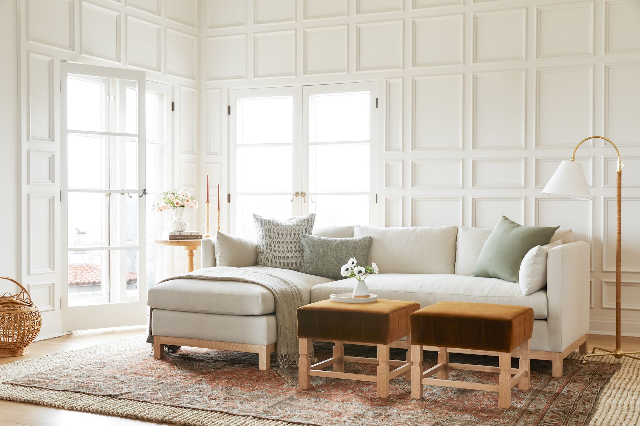 A bright living room featuring a large beige sectional sofa with grey and green throw pillows. Two brown ottomans are placed in front of the sofa on a patterned rug. A floor lamp and small round table with decor items are on the left side near large windows.