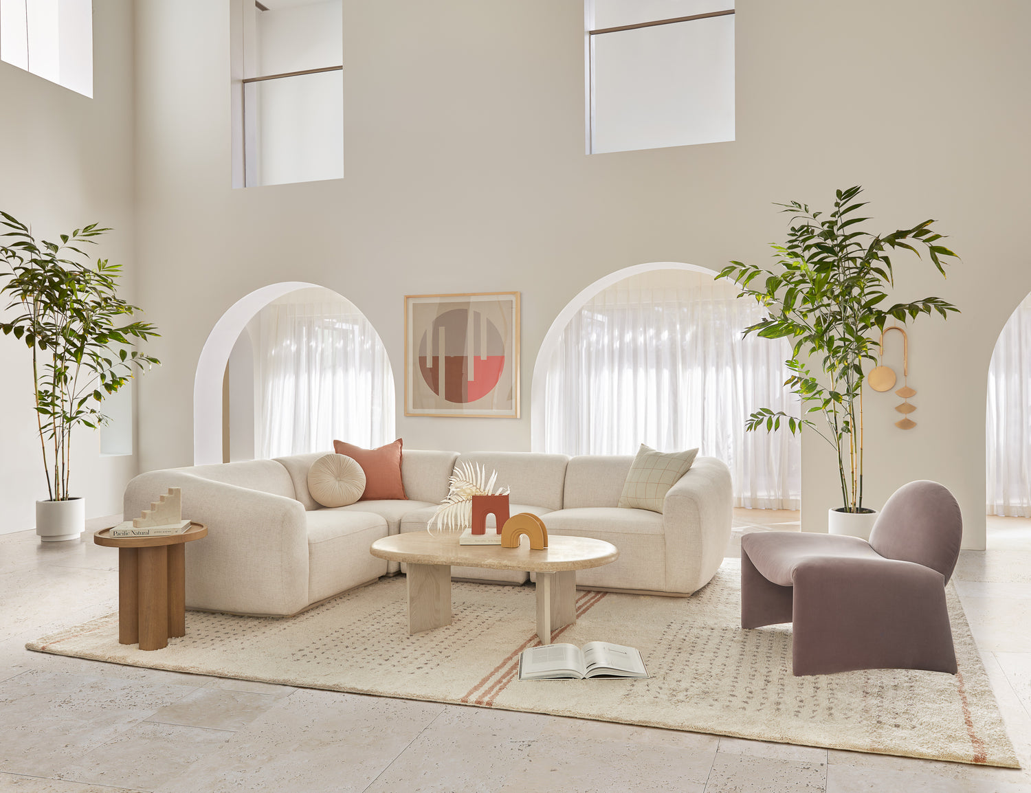 A spacious living room with high ceilings and large arched windows. It features a white sectional sofa, a round marble coffee table, and a plush armchair. Two potted plants flank the seating area, adding a touch of greenery. A modern artwork hangs on the wall.