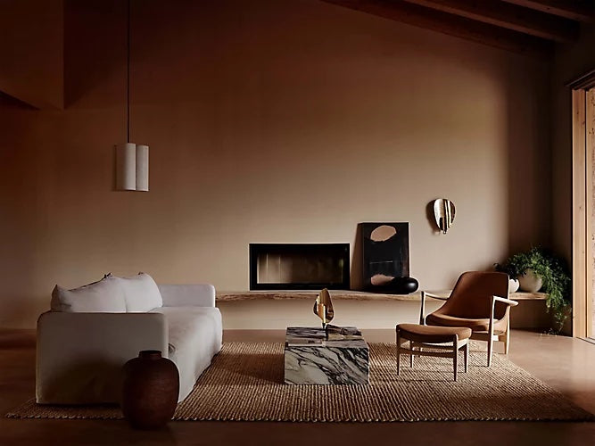 A minimalist living room with a beige color palette features a white sofa, a brown armchair, a marble coffee table, and a jute rug. A fireplace is set in the wall, and the room is decorated with hanging artwork, a ceramic vase, and a potted plant.