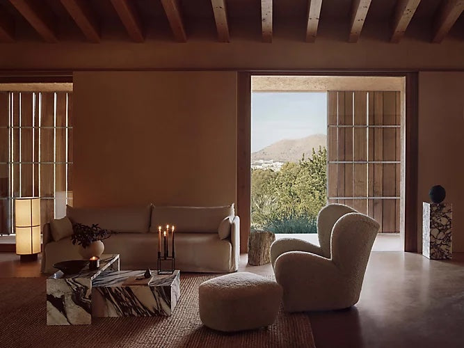 A cozy living room with natural light includes a white sofa, a plush armchair, a round ottoman, and a marble coffee table. Large sliding doors open to a view of greenery and mountains. Earthy tones dominate the decor with wooden ceiling beams and wicker rugs.