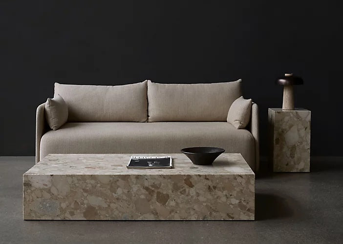 A minimalist living room setting features a beige sofa with two large cushions against a dark wall. In front, there is a rectangular stone coffee table holding a magazine and a black bowl. To the right, a matching stone side table holds a black lamp with a large shade.