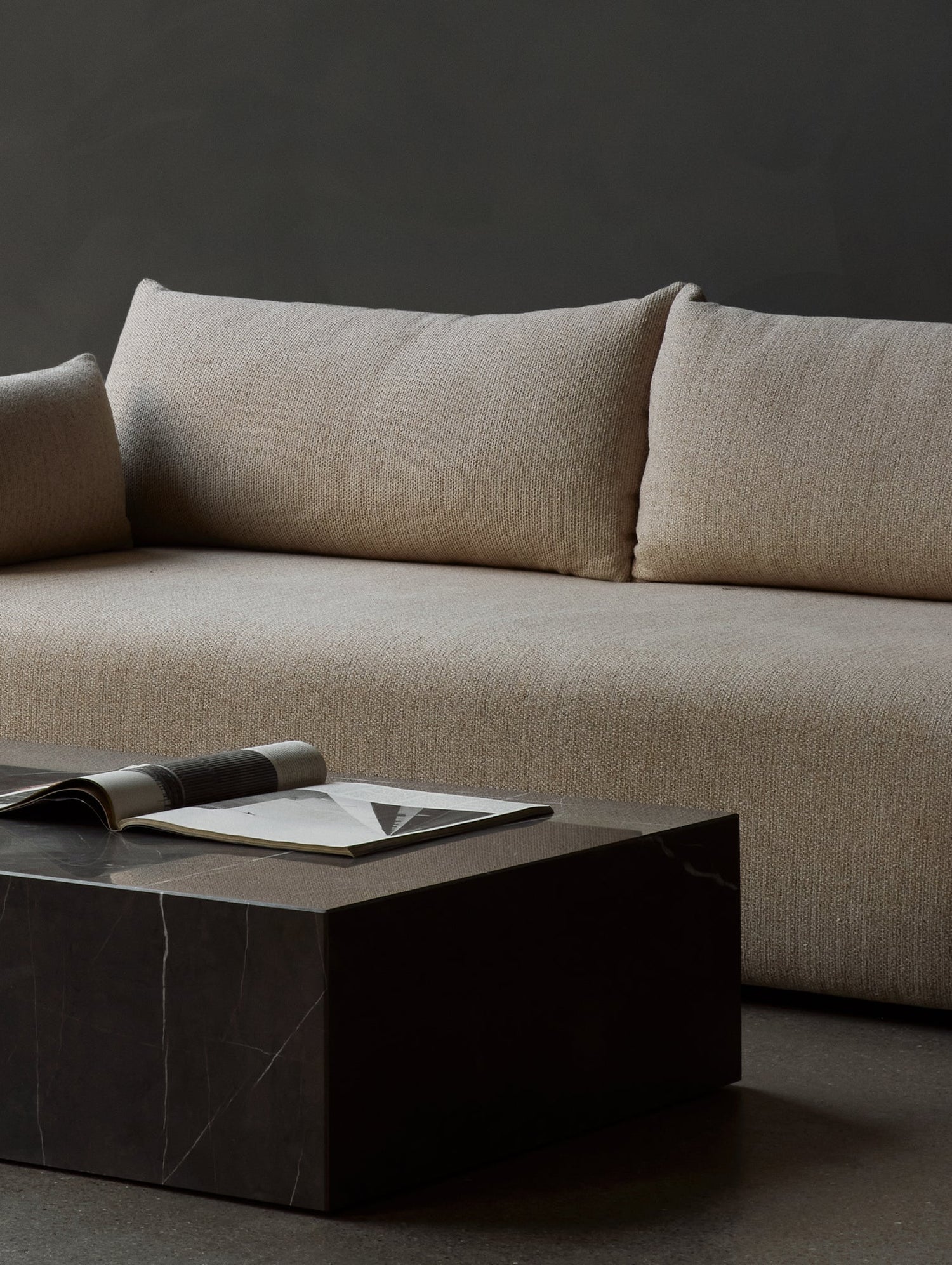 A modern beige sofa against a dark grey wall, accompanied by a sleek, dark marble coffee table. On the coffee table, there's an open book or magazine, showcasing a minimalist and contemporary living space.