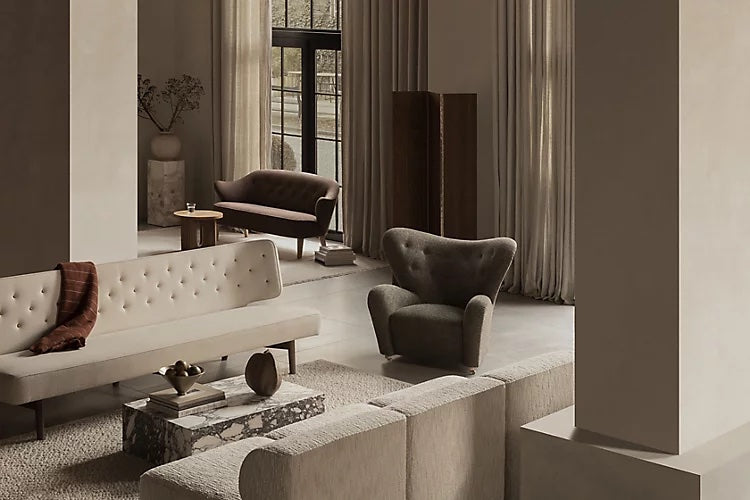 A contemporary living room with neutral tones features minimalist furniture including a beige sofa, a marble coffee table, a gray armchair, and a dark brown loveseat. Large windows with sheer curtains allow natural light to fill the space. A tall cabinet stands in the background.