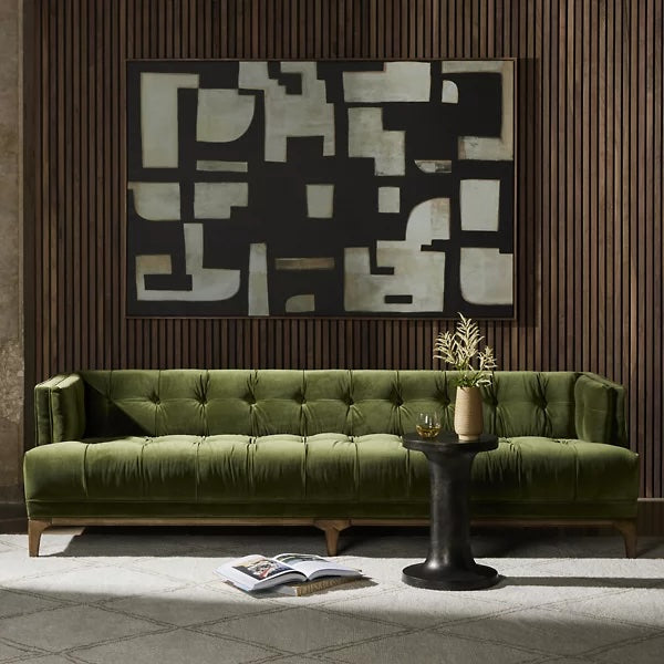 A modern living room with a deep green velvet tufted sofa, a small black side table holding a vase with flowers, and an abstract black and white geometric painting on a wood-paneled wall. The floor is carpeted in a light grey pattern.