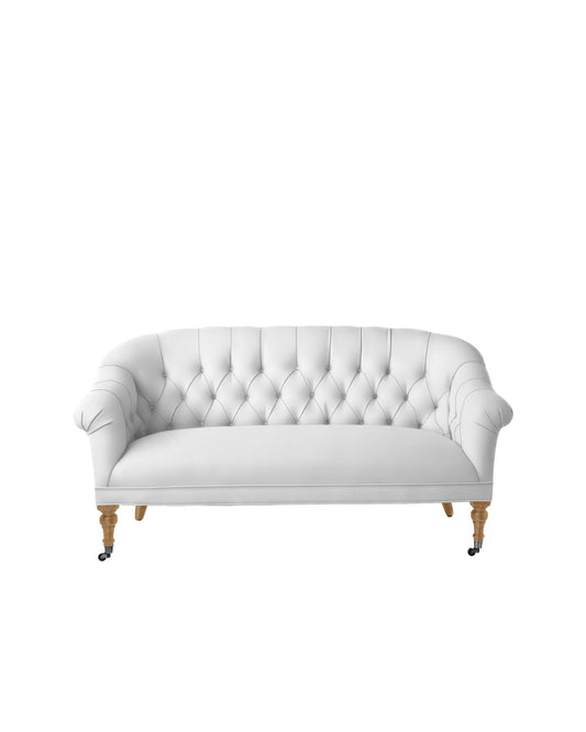 The Paxton Tufted Loveseat, featuring a white tufted design with rounded arms and wooden legs, including front wheels, is displayed against a plain white background.