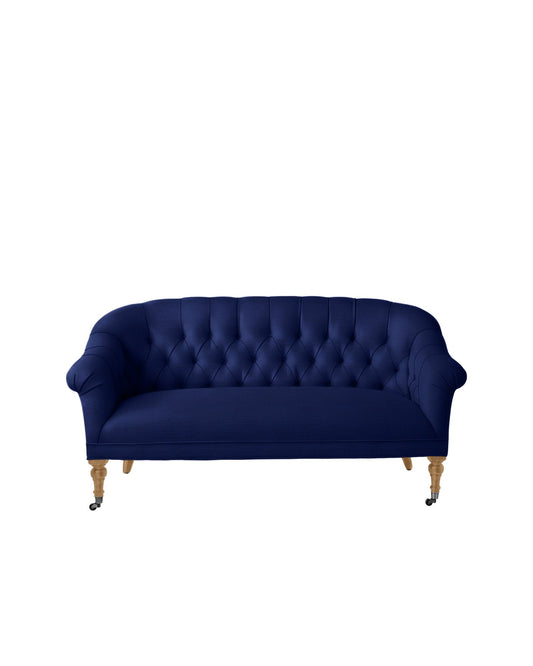 The Paxton Tufted Loveseat, in a rich deep blue color, showcases a curved back and wooden legs with small caster wheels. It is displayed against a plain white background.