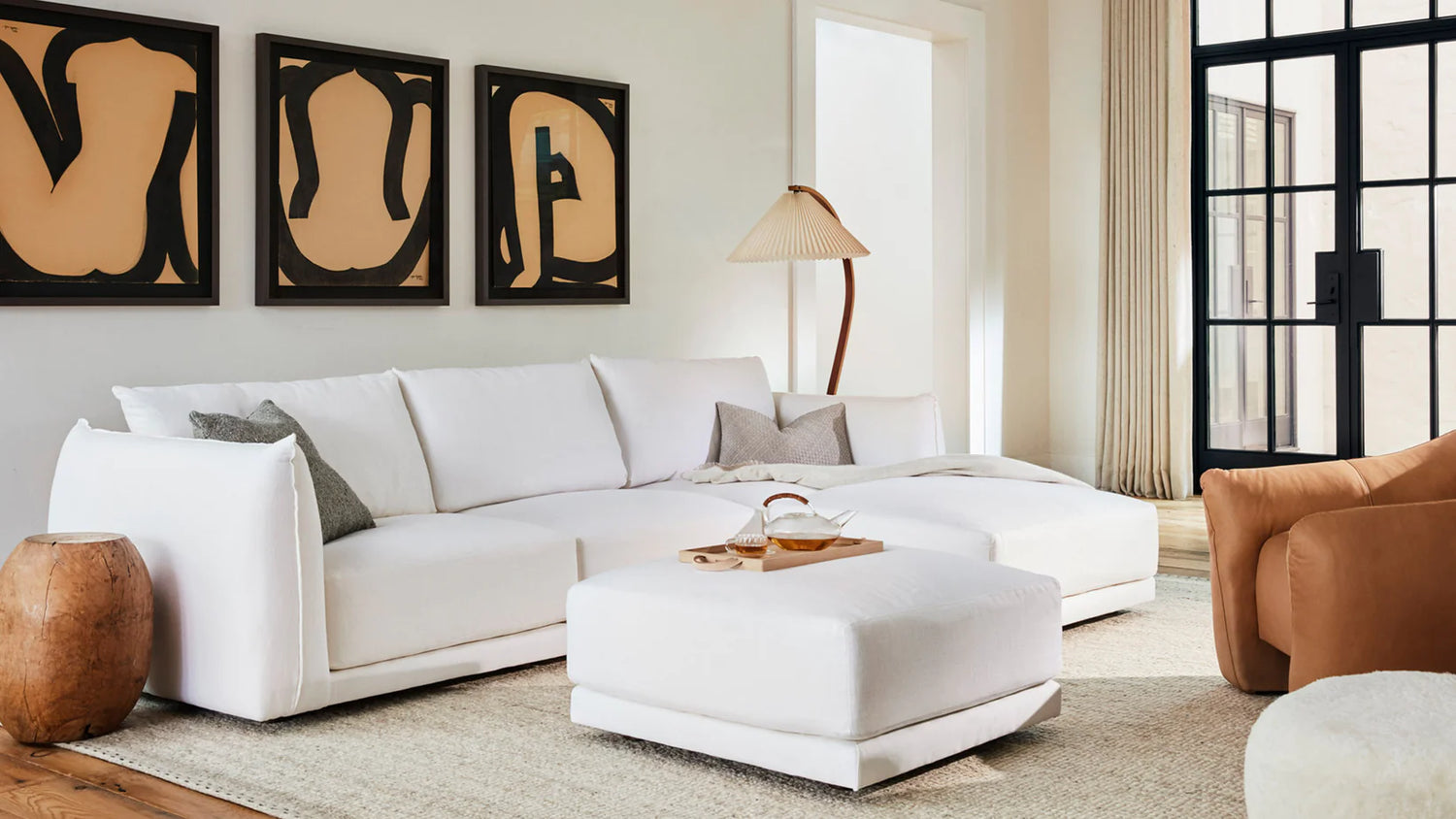 A modern living room with a white sectional sofa, white ottoman, and a wooden side table. Three abstract art pieces hang on the wall behind the sofa. A standing lamp with a beige shade is placed next to the sofa. Large windows allow natural light to fill the space.