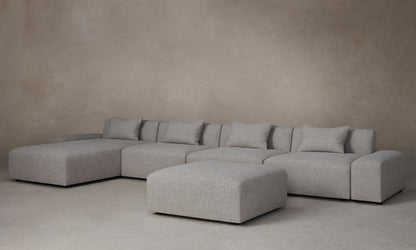 The Breuer Modular Chaise Armless Sectional 114" in Belgian Linen Agate, with its large, modern gray design and multiple cushions, is arranged against a plain beige wall. This sectional comprises various modular pieces, including an ottoman, offering a spacious and comfortable seating area.