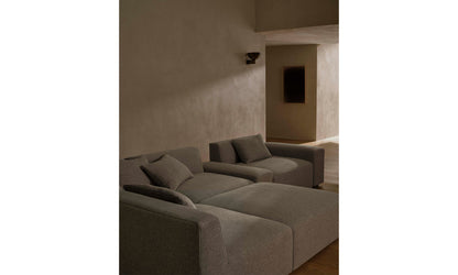 A minimalist living room showcases two large sofas from the Breuer Modular Chaise Armless Sectional 114" in Belgian Linen Agate, accented with soft cushions against a neutral-toned wall. A small wall light adds gentle illumination, fostering a cozy atmosphere, while a dark, rectangular wall decoration subtly adorns the background.