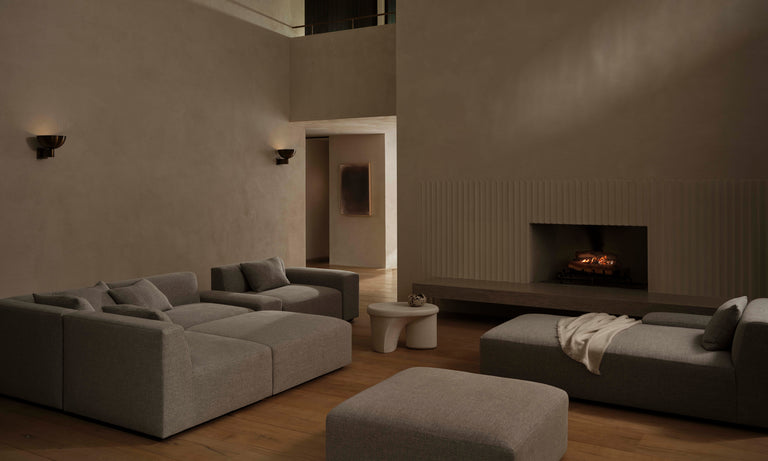 A contemporary living room featuring The Breuer Modular Chaise Armless Sectional 114" in Belgian Linen Agate, complemented with matching cushions and a square ottoman, alongside a minimalist white coffee table. A large fireplace is to the right, with a cozy blanket draped over the sectional's front. The room is gently illuminated by wall sconces.