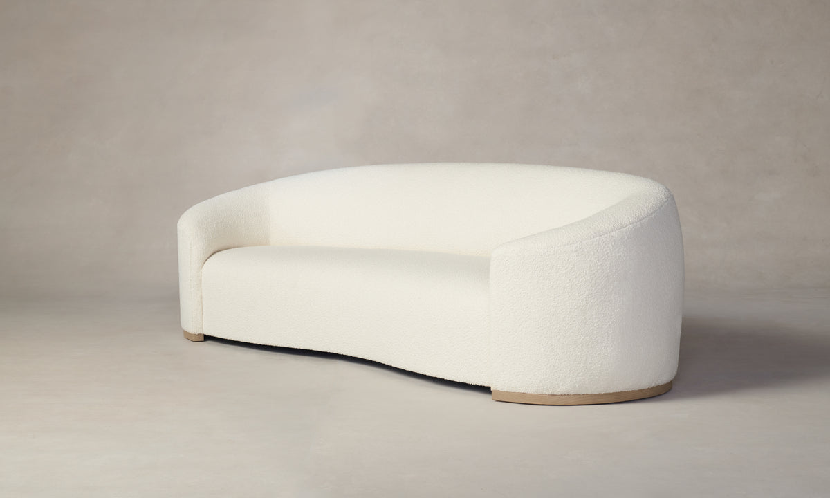 The Chelsea Sofa in Italian Bouclé White features a modern, crescent shape with a textured fabric finish. This sofa boasts a smooth, minimalist design with curved edges and a light wood base, elegantly complemented by an understated beige background.