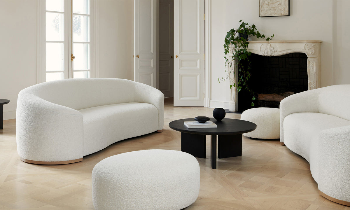 A contemporary living room showcasing The Chelsea Sofa in Italian Bouclé White, elegantly curved and complemented by an ottoman on a light wood floor. In the center sits a sleek black circular coffee table. A fireplace adds warmth to the room, with a plant nearby set against pristine white walls and double doors.