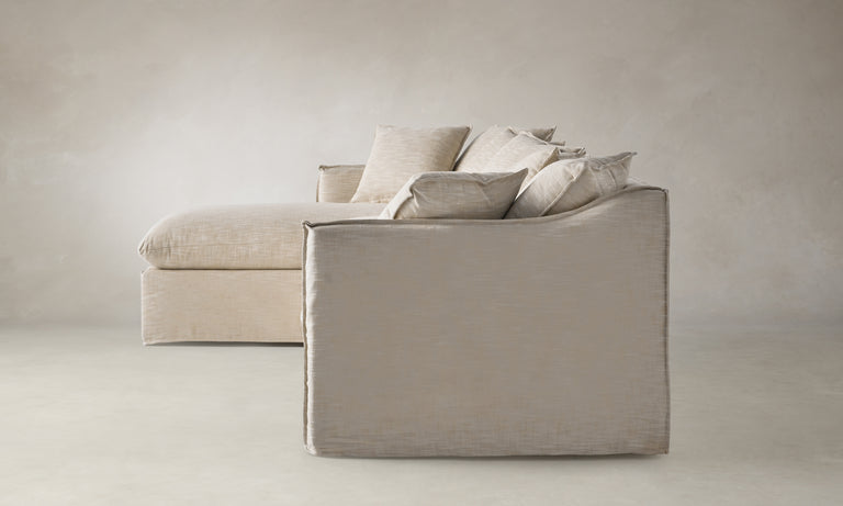 The Dune 90" Sectional in Belgian Linen Alder is placed on a light-colored floor against a neutral wall. This beige sofa showcases plush seat cushions and numerous pillows, crafting a cozy and inviting atmosphere.