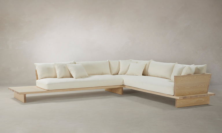 The Muir Sectional, a modern L-shaped sofa with a light wood frame and off-white cushions, is set against a neutral background. It features multiple throw pillows and an integrated low wooden side table.