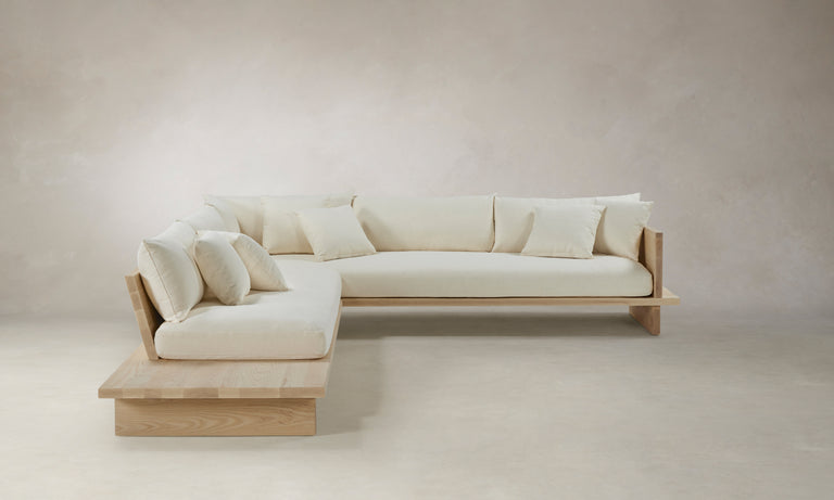 The Muir Sectional, an L-shaped sofa featuring cream-colored cushions and a wooden frame, stands against a neutral-colored wall. Its modern and clean design emphasizes simplicity and natural materials.