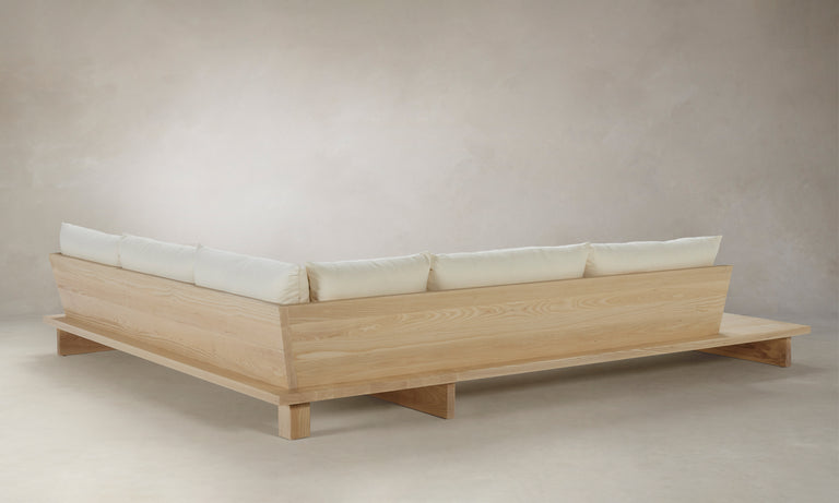 The Muir Sectional is a minimalist, light wooden platform bed with off-white cushions arranged against a plain background. It features clean lines and a modern, low-profile design.