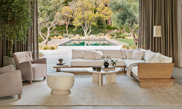 A spacious living room opens to a tranquil outdoor pool area and is furnished with The Muir Sectional in beige, two armchairs, a round pouf, and a wooden coffee table. Large windows with curtains allow natural light to flood the space, enhancing views of the lush greenery outside.