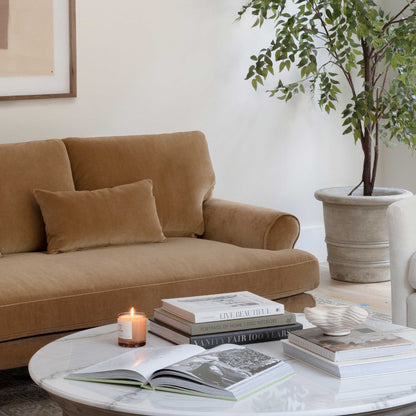 A cozy living room featuring the Maxwell Custom Sofa in a tan hue, complemented by a round marble coffee table adorned with stacked books, a lit candle, and a decorative bowl. A large potted plant and an art piece on the wall enhance the space with greenery and style.