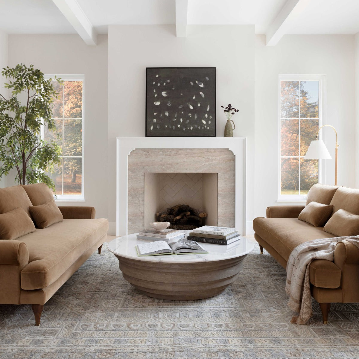 A cozy living room featuring two Maxwell Custom Sofas facing each other, a round marble coffee table at the center, and a fireplace adorned with a black and white abstract painting above it. Large windows on either side allow natural light to stream in.