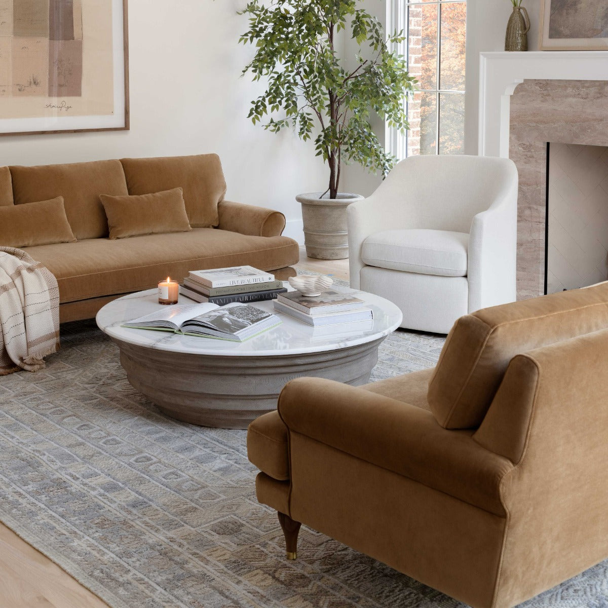 A cozy living room featuring the Maxwell Custom Sofa in a warm brown shade, complemented by a white armchair. The room is arranged with a round coffee table displaying books and a candle, laid over a patterned rug. A potted plant sits by the window, and the décor exudes warmth and invitation as natural light fills the space.