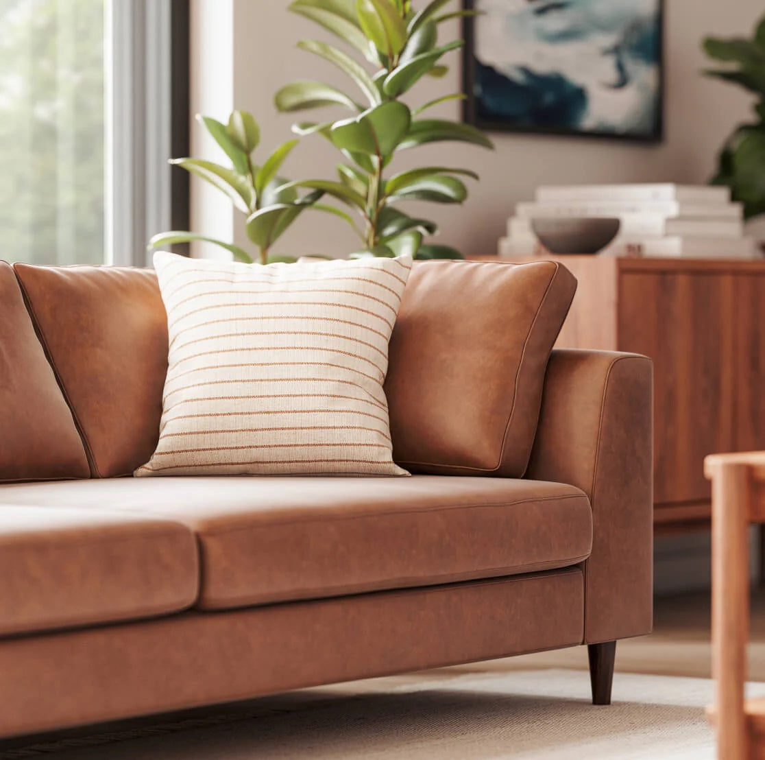 A modern living room features a brown leather sofa with a white and brown striped pillow. A potted plant sits beside the sofa, and a framed abstract artwork hangs on the wall. A wooden cabinet with books adds a cozy touch to the space.